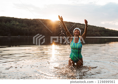Cold water swimming for elderly women. Senior sporty women splashing in the water, standing in lake during cold evening, arms in the air. 119899561