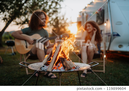 Couple sitting by campfire in the evening, enjoying peaceful moment. Man playing guitar and singing song to his girlfriend. Camping trip for young people. 119899604