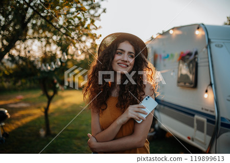 Portrait of young beautiful woman on a camping trip in nature, standing in front of caravan with smartphone in hand. 119899613