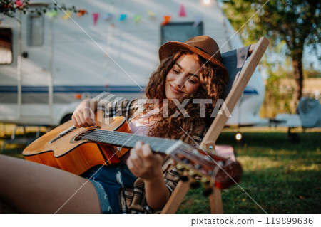 Young beautiful woman is on a camping trip in nature, sitting in front caravan and playing guitar. 119899636