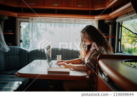 Young beautiful woman is on a camping trip in nature, sitting in her caravan and working on a notebook. Caravan as homeoffice, working remotely. 119899644