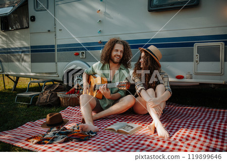 Couple sitting by caravan, enjoying peaceful moment. Man playing guitar and singing song to his girlfriend. Camping trip for young people. Couple sitting by caravan, enjoying peaceful moment. Man playing guitar and singing song to his girlfriend. Camping trip for young people. 119899646