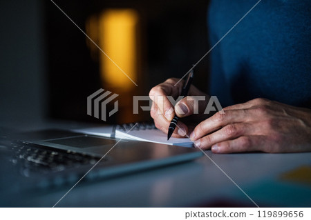 Close up of man working from home office at evening, writing notes in planner. Telecommuting and home office. 119899656