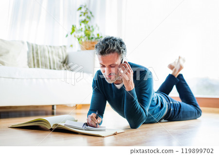 Portrait of handsome man lying on belly and making phone call, writing notes in planner. 119899702