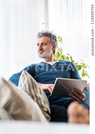 Portrait of attractive middle aged man at home, watching tv show on tablet, sitting on sofa in living room. 119899704