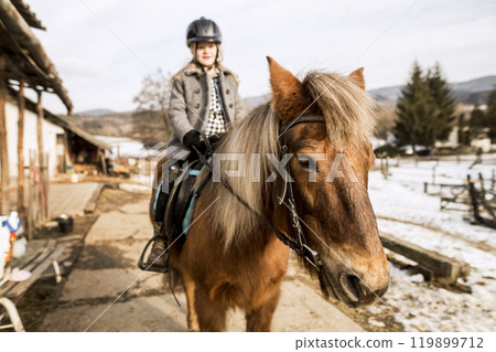 Young girl spending cold winter weekend on family farm, riding her brown fluffy pony. Young girl spending cold winter weekend on family farm, riding her brown fluffy pony. 119899712
