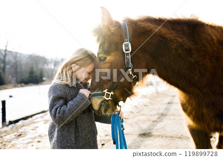 Young girl spending cold winter weekend on family farm, taking care of her brown fluffy pony, hugging him. 119899728