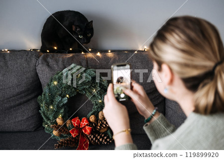 Woman photographing black cat with Christmas wreath and lights, creating user-generated holiday content. Cozy home setting with festive decorations. 119899920