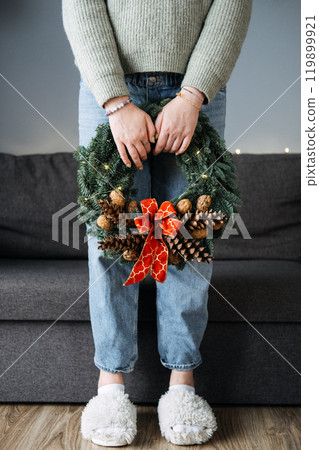 Person holding handmade Christmas wreath with pinecones and red bow, standing in cozy home setting. DIY holiday decor, festive minimalism, and winter crafts. Person holding handmade Christmas wreath with pinecones and red bow, standing in cozy home setting. DIY holiday decor, festive minimalism, and winter crafts. 119899921