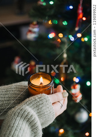 Hands holding a lit candle in cozy sweater, making a wish for Christmas. Warm and festive holiday atmosphere with Christmas tree in background. Hands holding a lit candle in cozy sweater, making a wish for Christmas. Warm and festive holiday atmosphere with Christmas tree in background. 119899922