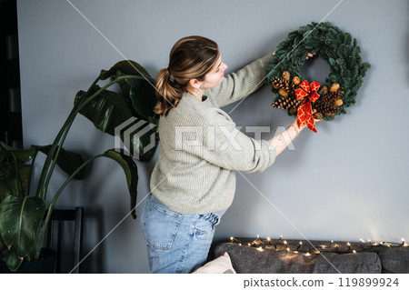 Woman hanging handmade Christmas wreath with pinecones and red ribbon. DIY holiday decor adds a personal touch to festive home decorating. 119899924