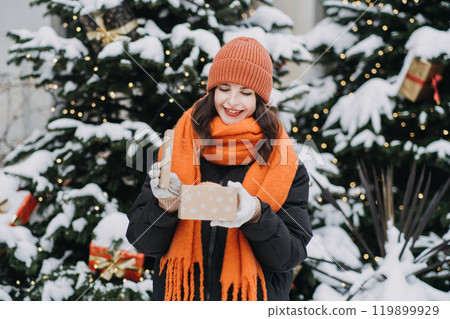 Woman with orange scarf unboxing a gift in a snowy Christmas scene. Festive outdoor gift-giving and holiday celebration in cozy winter fashion 119899929