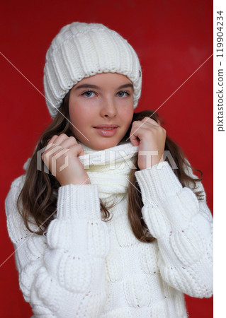 a young woman with long chestnut hair is playfully obscured by a cozy white knitted hat that rests over her eyes. She holds a soft, textured scarf in her hands against a vivid red background.. 119904234