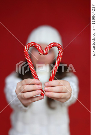 A cheerful young woman with long chestnut hair wears a cozy white knitted hat and scarf, holding colorful lollipops against a vibrant red background. 119904237