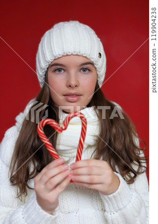 A cheerful young woman with long chestnut hair wears a cozy white knitted hat and scarf, holding colorful lollipops against a vibrant red background. 119904238