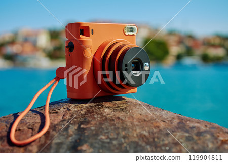 Vintage orange instant camera on rock by seaside with blurred coastal town in background. Summer travel concept. Equipment for capturing moments 119904811