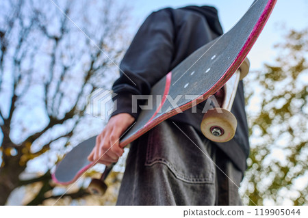 Skateboarder holding worn skateboard walks at street Skateboarder holding worn skateboard walks at street 119905044