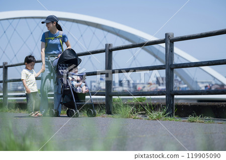 一位母親和她的兄弟姊妹(一家三口)推著嬰兒車在陽光明媚的河濱公園散步,手牽手進行春夏郊遊 一位母親和她的兄弟姊妹(一家三口)推著嬰兒車在陽光明媚的河濱公園散步,手牽手進行春夏郊遊 119905090