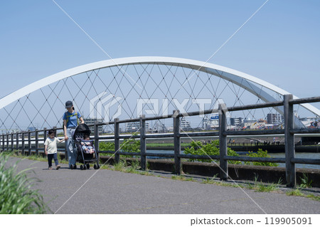 一位母親和她的兄弟姊妹（一家三口）推著嬰兒車在陽光明媚的河濱公園散步，手牽手進行春夏郊遊 119905091