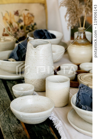 Rustic table setting with empty craft handmade ceramic tableware, white rough bowls, plates, cups, jug and vase on linen tablecloth. Dry reed flowers. Day light dining room. 119906143