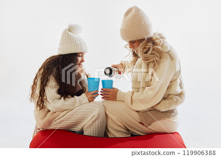 Mother pouring hot drink for her daughter from thermos as they sit on snow tube, taking break from dynamic winter activities. Mother pouring hot drink for her daughter from thermos as they sit on snow tube, taking break from dynamic winter activities. 119906388