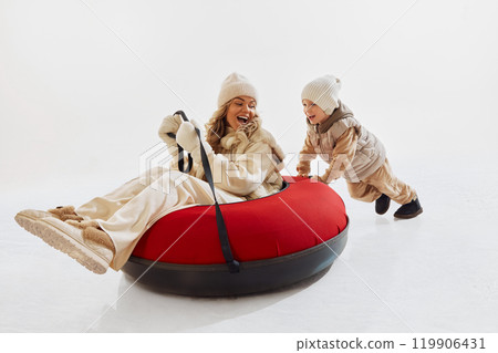 Cozy happy moment between mother and son. Happy mother sitting on red snow tube and little boy pushing it forward Cozy happy moment between mother and son. Happy mother sitting on red snow tube and little boy pushing it forward 119906431