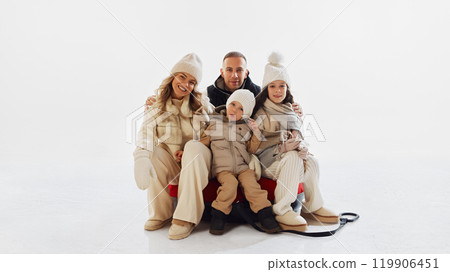 Portrait of family, mother, father and little boy and girl, dressed in stylish warm clothes spending day together, sitting on red snow tube. 119906451