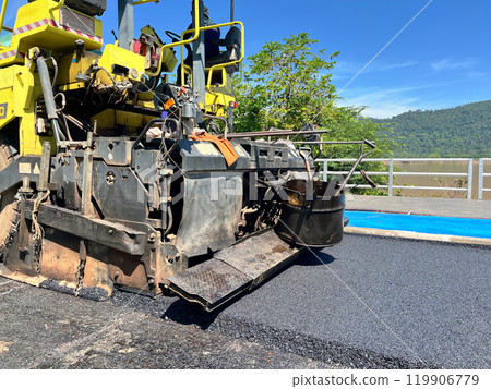 asphalt paver machine laying fresh asphalt on a road. The image captures the machinery in action, showing the spreader screed and other components, smooth layer of asphalt, construction site, civil 119906779