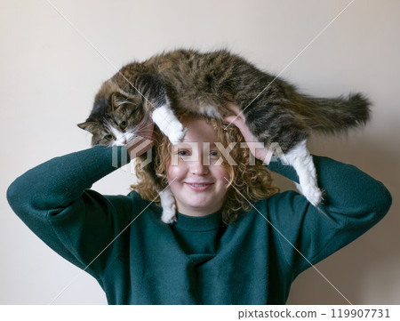 Girl with curly hair smiles widely while holding her arms up, supporting a cat resting atop young adult's head. The setting is warm and inviting, capturing a fun moment between them. Light background. 119907731