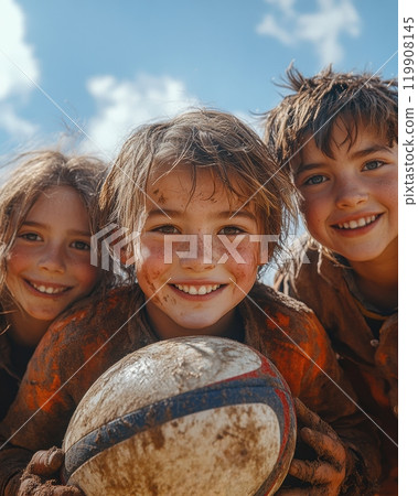 Three happy children with muddy faces holding rugby ball, enjoying outdoor game. 119908145