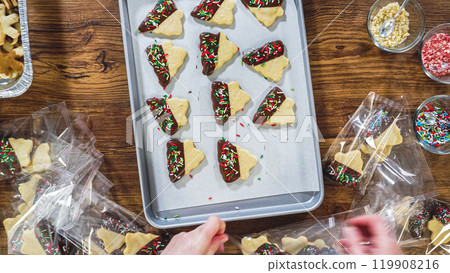 Flat lay. Carefully packaging Christmas cutout cookies, half-dipped in chocolate, sprinkled with crushed nuts, and presented in clear cellophane wrapping. Flat lay. Carefully packaging Christmas cutout cookies, half-dipped in chocolate, sprinkled with crushed nuts, and presented in clear cellophane wrapping. 119908216