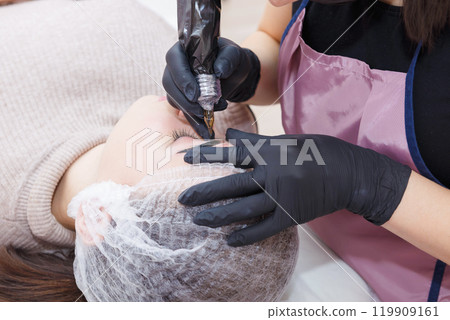 The master applies eyebrow makeup paint to the client with a tattoo machine. The master applies eyebrow makeup paint to the client with a tattoo machine. 119909161