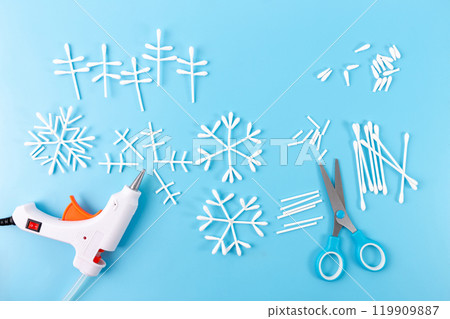 Handmade cotton swab snowflake decorations on blue background step in creative process, with hot glue gun and scissors beside. DIY craft idea using everyday materials. 119909887
