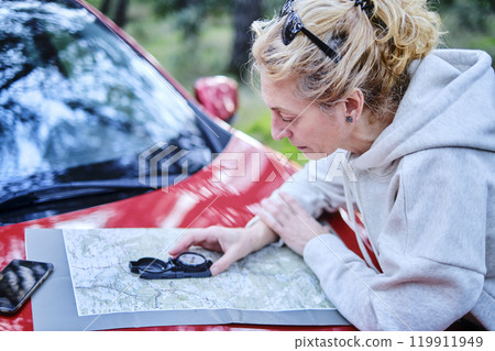 Woman orienting herself with a map and a compass close to a car in a forest. 119911949