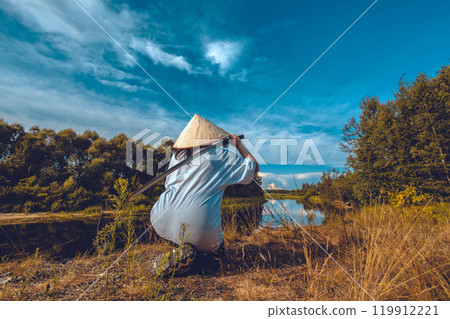 A samurai warrior in traditional attire kneels by the river, holding his katana at the ready, under a blue sky, radiating calm and strength. A samurai with a katana kneels on the bank of a river. 119912221