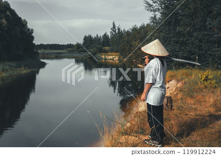 Samurai with katana in thought. A young samurai in traditional clothing and a conical hat stands by a river holding a katana, looking at a serene landscape, representing peace and tranquility. Samurai with katana in thought. A young samurai in traditional clothing and a conical hat stands by a river holding a katana, looking at a serene landscape, representing peace and tranquility. 119912224