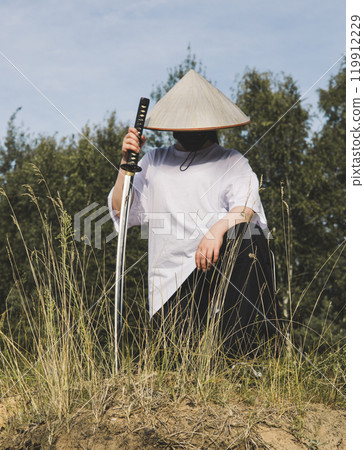 A warrior katana on the river bank. A man dressed as a samurai, with a conical hat and mask, kneels in nature holding a katana, creating a striking contrast between tradition and modernity. 119912229