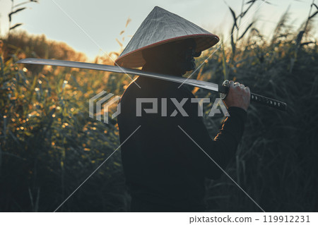 A samurai warrior in a conical hat and black clothes holds a katana on his shoulder at sunset in a field of tall grass. A handsome samurai looks into the distance with a katana on his shoulder. 119912231