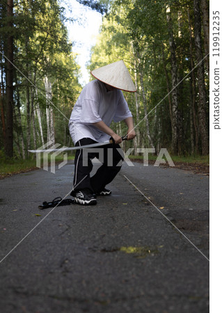 A focused samurai wearing an Asian hat practices with a katana in the forest embodying Japanese culture with skill and strength. The stance exudes the balance and strength of a warrior with a katana. 119912235