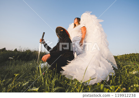 A masked samurai crouches in a field holding a katana next to an angel with large white wings, creating a striking juxtaposition against the sunset. Samurai and angel together at sunset. 119912238