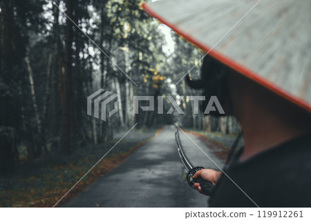 A samurai warrior in a straw hat holds a katana while walking along a forest road. The image evokes a sense of mystery and adventure. The samurai, with a katana in his hands, is ready to fight. A samurai warrior in a straw hat holds a katana while walking along a forest road. The image evokes a sense of mystery and adventure. The samurai, with a katana in his hands, is ready to fight. 119912261