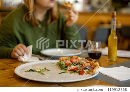A woman holds a piece of pizza in hands. Focus on a pizza. Pizza with prosciutto, ham, arugula, tomatoes and parmesan. Italian cuisine. Food banner A woman holds a piece of pizza in hands. Focus on a pizza. Pizza with prosciutto, ham, arugula, tomatoes and parmesan. Italian cuisine. Food banner 119912868