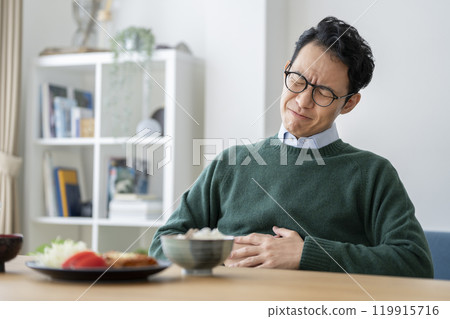 一名 40 歲左右的男子在餐桌上把手放在肚子上，出現腹痛、沉重、食慾不振的圖片 119915716