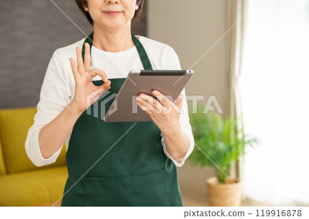 A senior woman housewife/housekeeper wearing a green apron looking at a tablet in the living room at home and making a circle sign A senior woman housewife/housekeeper wearing a green apron looking at a tablet in the living room at home and making a circle sign 119916878