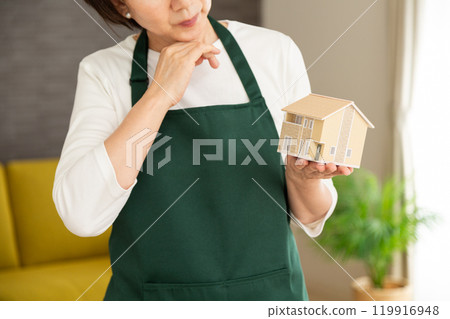 一位身穿綠色圍裙的高級女主婦/管家在自家客廳裡拿著房屋模型，憂心忡忡。 119916948