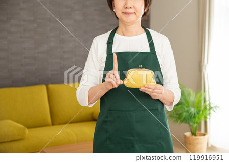 A senior female housewife/housekeeper wearing a green apron holding a purse and pointing her index finger up in the living room at home 119916951