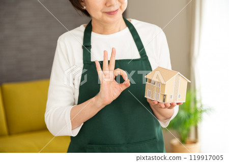 一位身穿綠色圍裙的高級女主婦/管家拿著房屋模型,在她家的客廳裡做一個圓圈標誌 一位身穿綠色圍裙的高級女主婦/管家拿著房屋模型,在她家的客廳裡做一個圓圈標誌 119917005