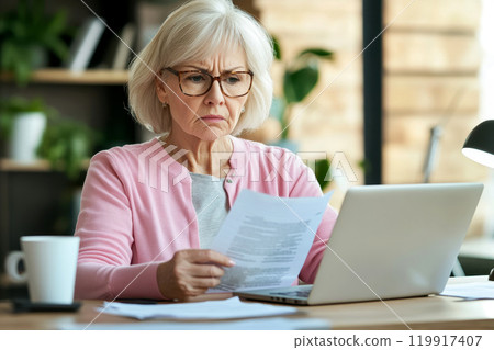 Older woman with serious expression read document sitting at desk with laptop, dealing with important paperwork, managing finances or health-related issues, learn written information looks concerned 119917407