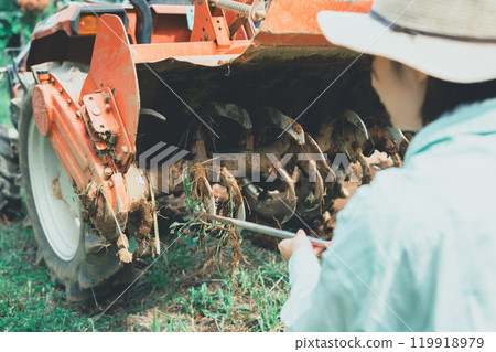 A young woman removing mud from a tractor rotary 119918979