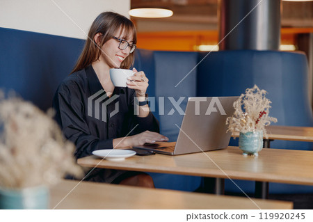 A Young Woman Engaged in Work on Her Laptop While Enjoying a Delicious Coffee at a Modern Cafe A Young Woman Engaged in Work on Her Laptop While Enjoying a Delicious Coffee at a Modern Cafe 119920245